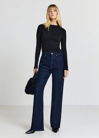 Schwarzes, geripptes Langarmshirt kombiniert mit hochtaillierten, dunkelblauen Jeans mit weitem Bein und dezenten Verzierungen sowie einer schwarzen Pelzclutch.