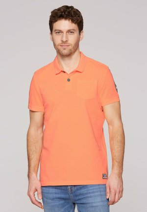 Hombre con cabello rizado y castaño, vestido con una camisa polo naranja brillante y vaqueros azules, de pie frente a un fondo gris liso.