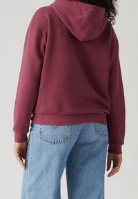 Persona con un hoodie color burdeos con la capucha hacia abajo y jeans de mezclilla azul claros, mostrado desde detrás contra un fondo liso.