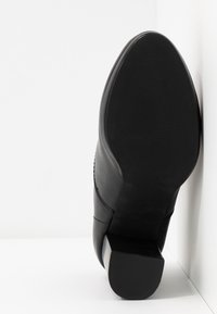 Chaussure en cuir noir à bout pointu et talon bloc vue par la semelle, appuyée contre un angle mur et sol blanc.