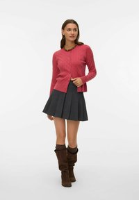 Roza kardigan iz pletenega bombaža z gumbi spredaj, kombiniran z nagubano sivo krilo in rjavimi ohlapnimi škornji do kolen. Videz dopolnjuje ogrlica.