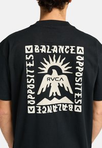 T-shirt di cotone nero con una grande grafica bianca con il testo "BILANCIARE GLI OPPOSTI" e un'immagine di un sole e un uccello, centrata sul retro.