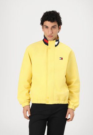 Chaqueta amarilla hecha de tejido ligero, con un alto cuello adornado con franjas rojas, blancas y azules. Puños y cinturilla elásticos. Pequeño logo en el pecho.