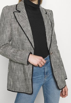 Kvinde iført sort-hvid hundetand mønstret blazer med sort kant, sort højhalset trøje og blå jeans, hånd hvilende ved taljen.