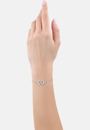 Bracciale d'argento con un ciondolo a forma di cuore decorato con pietre ornamentali, montato su una delicata catena. Mano mostrata in posizione sollevata.