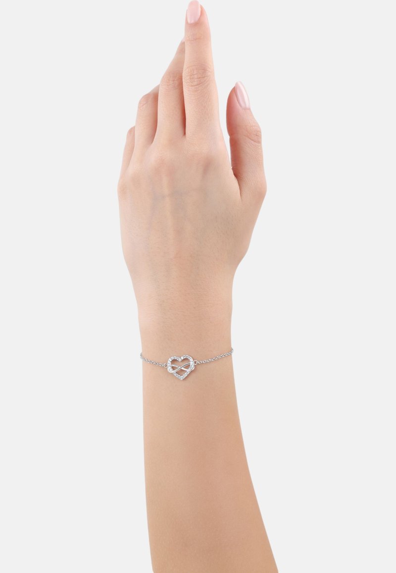Pulsera de plata con un charm en forma de corazón adornado con piedras decorativas, montada en una delicada cadena. Mano mostrada en una posición elevada.