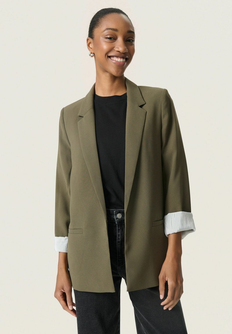 Blazer verde măsliniu cu un design structurat, având manșete cu dungi răsucite și un croi lejer. Purtați peste un top negru și blugi închiși la culoare.