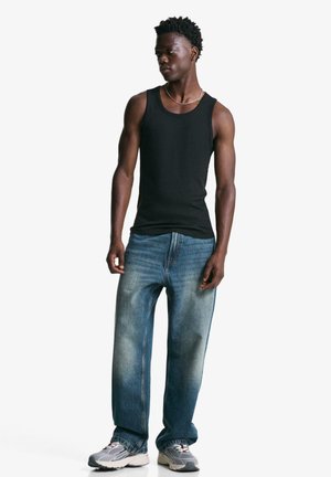 Schwarzes Tanktop kombiniert mit ausgebleichten blauen geraden Jeans. Model trägt graue Sneakers und steht vor einem weißen Hintergrund.