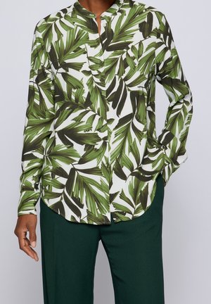 Persoon draagt een wit overhemd met lange mouwen met groene palmbladprint en donkergroene broek, staand met één hand in de zak.