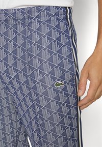 Blå mönstrade sweatpants med geometrisk design i vitt, elastisk midja och en grön krokodil-logga. Sidobanor i kontrasterande färg.