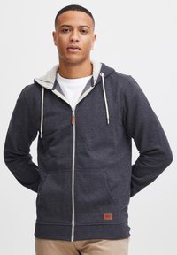 Sudadera de cremallera en color carbón con capucha forrada de felpa blanca, cordones, dos bolsillos frontales y una etiqueta de cuero marrón en el dobladillo inferior.