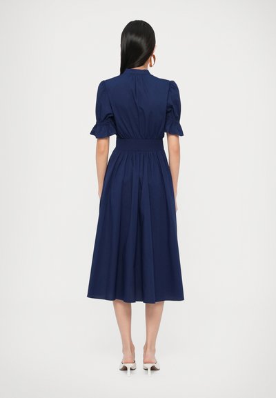 Φόρεμα σε ναυτικό μπλε με φουσκωτά μανίκια, εφαρμοστή μέση και αέρινο μήκος μέχρι το μέσο της γάμπας. Λείο ύφασμα με λεπτομέρειες σε φουσκωτή φούστα.