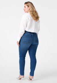 Chemise blanche à boutons, ornée de clous argentés, associée à un jean skinny taille haute bleu et des escarpins argentés, photographiée de dos.