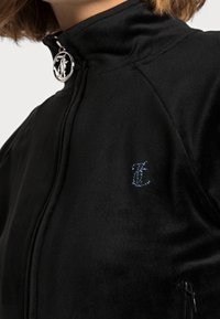 Black velvet zip-up jacket featuring a high collar, shiny silver hardware, and a blue embroidered logo on the chest. Soft texture with fitted design.