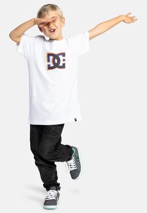Witte T-shirt met korte mouwen en een kleurrijk DC-logo, gecombineerd met zwarte denim jeans en grijze sneakers met mintaccenten.