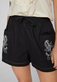 Calções pretos com um cós elástico e cordão. Apresentam bordados florais brancos nos bolsos laterais e costura em contraste.