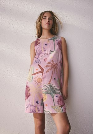 Ung kvinde iført lyserød ærmeløs kjole med tropiske print, stående mod en struktureret hvid væg og kiggede let opad.