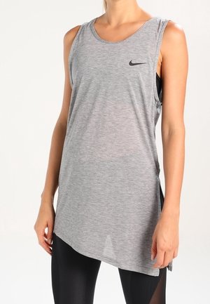 Femme portant un débardeur gris sans manches Nike avec un logo noir, assorti à un legging noir, debout devant un fond clair uni.