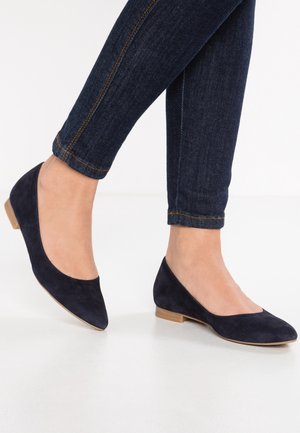 Marineblå ruskinds flade sko med spidse tæer, lav hæl og glat overflade; parret med mørk denim, der fremhæver ankeldetaljer.