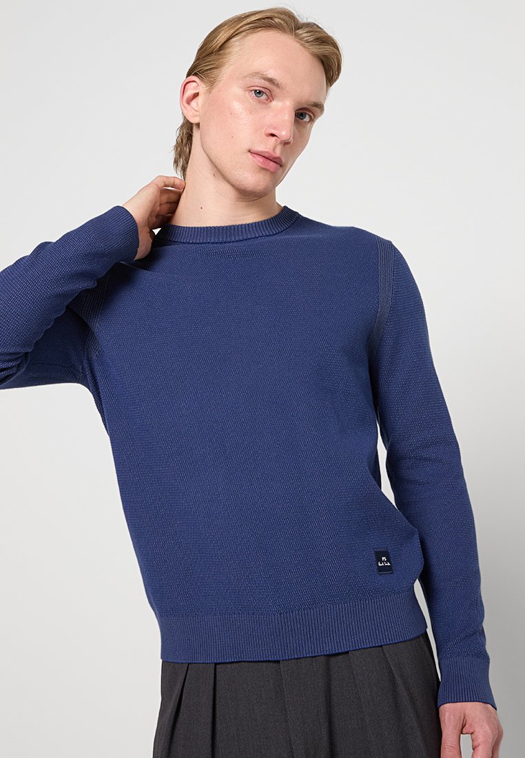 Ung person med ljust hår som bär en blå stickad tröja och mörkgrå byxor, poserar med en hand bakom nacken.