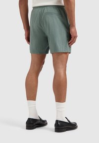 Groene, textuurshorts met een elastische tailleband, voorzien van een achterzak. Draagt zwarte schoenen en witte geribbelde sokken.