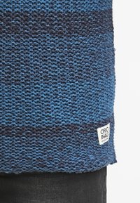 Sweter z dzianiny w różnych odcieniach niebieskiego z teksturowanym wzorem. Posiada małą białą metkę z napisem „ORIGINALS” przy dolnym brzegu.