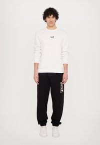 Hvid sweatshirt med rund hals og lille sort logo. Sorte joggingbukser med trykt logo i cremefarve. Hvide sneakers fuldender outfittet.