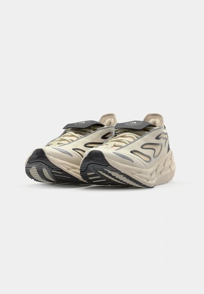 Zapatillas deportivas de color beige claro con una parte superior texturizada, detalles en negro y un diseño ondulado. Cuentan con una suela gruesa y un patrón de relieve intrincado.
