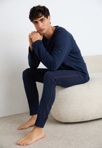 Navy-blaues Langarmoberteil und Hosen-Set aus glattem Material mit roten Seitestreifen. Modell sitzt auf einem strukturierten beigen Sitz und Teppich.