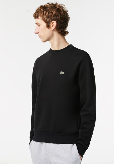 Sweat-shirt noir avec col et poignets en rib, arborant un petit logo crocodile vert de Lacoste sur la poitrine. Texture lisse et coupe classique.