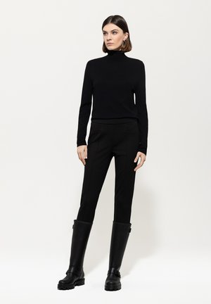 Woman wearing a black turtleneck, slim black pants, and knee-high black boots, standing against a plain light background.
