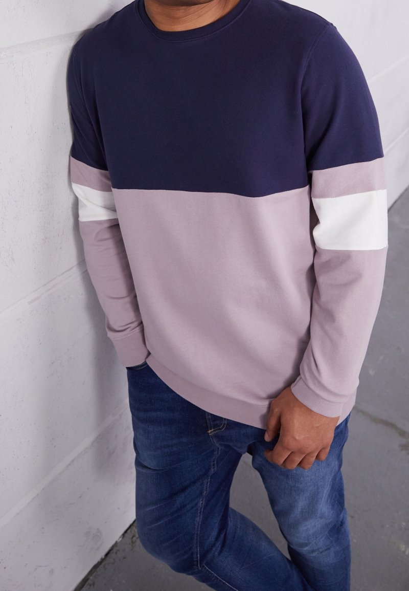 Homme portant un sweat-shirt bicolore avec des parties bleu marine, lavande et blanc, associé à un jean bleu, debout contre un mur clair.
