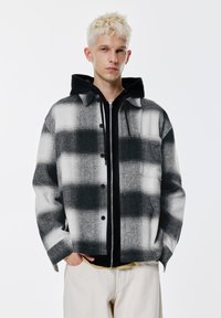 Crno-bijela karirana jakna s prednjim zakopčavanjem na gumbe, teksturiranim tkanjem i crnom kapuljačom ispod. Izgled zaokružuju slonovače hlače.