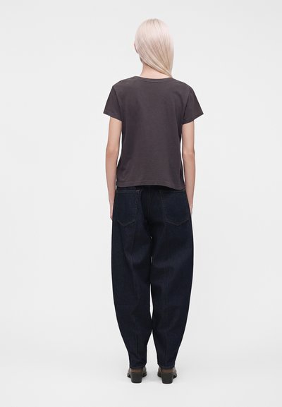 Personne aux longs cheveux blonds portant un t-shirt gris foncé à manches courtes, un jean bleu foncé ample et des chaussures noires, debout de dos.