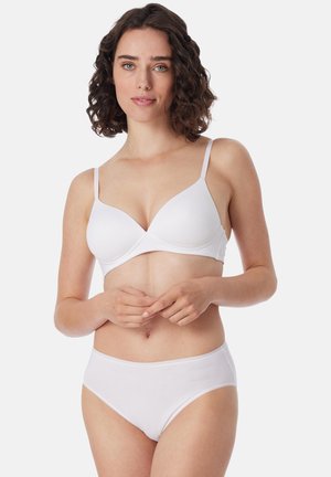 Femme aux cheveux courts et bouclés portant un soutien-gorge blanc et un slip, debout devant un fond clair uni, les mains jointes devant elle.