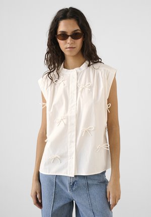 Frau trägt ärmellose weiße Bluse mit kleinen Schleifen, blaue Jeans und braune Sonnenbrille, steht vor einfarbigem grauem Hintergrund.