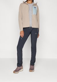 Nainen, joka on pukeutunut beigeen vetoketjulliseen takkiin, jossa on vaaleansininen rintatasku, harmaaseen t-paitaan, mustiin slim-fit-housuihin ja mauvenvärisiin vaelluskengiin, seisoo.