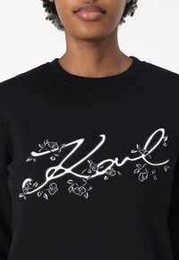 Sudadera negra con cuello redondo que presenta texto bordado en blanco "Karl" y diseños florales en el frente. Textura de tela suave.