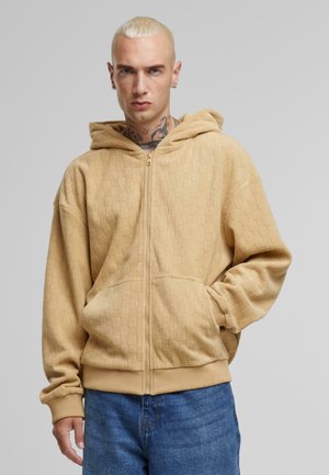 Felpa con cappuccio beige, caratterizzata da un pattern a griglia testurizzato, chiusura con zip e tasche frontali. Polsini e orlo a costine, tessuto morbido.