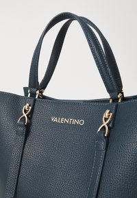 Modra strukturirana usnjena torbica z zlatimi kovinskimi elementi, dvojni ročaji in izrazitimi blagovnimi imeni na sprednji strani.