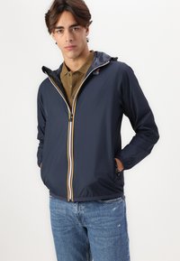 Navy lightweight jacket with contrasting yellow and white zipper, side pockets, adjustable hood, and elastic cuffs. Worn over a brown polo shirt.