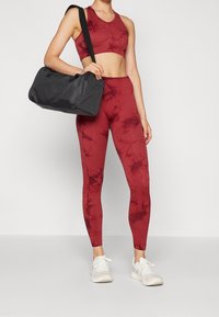 Czerwony zestaw sportowy z topem w stylu tie-dye i legginsami oraz czarną torbą na siłownię. Legginsy są dopasowane, a torba ma długi pasek i zamek błyskawiczny.