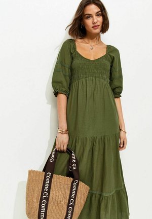 Mujer con vestido verde de volantes y mangas abullonadas, sosteniendo un bolso tote tejido con asas negras etiquetado "Comme Ci, Comme Ça".