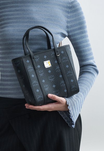 Frau in blau gestreiftem Oberteil und schwarzen Nadelstreifen-Hosen hält eine kleine schwarze Ledertasche von MCM mit Logo-Muster und silberner Platte.