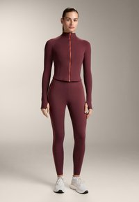 Bordó, testhez simuló sportos szett, hosszú ujjakkal, narancssárga cipzárral és oldalsávokkal. Az anyag simának és rugalmasnak tűnik, edzéshez alkalmas.