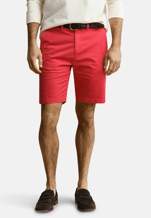 Mann trägt leuchtend rote Shorts, schwarzen geflochtenen Gürtel, cremefarbenes Langarmshirt und dunkelbraune Loafers vor einfachem Hintergrund.