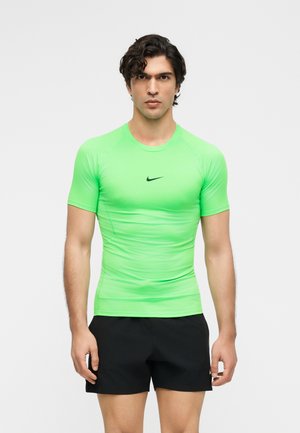 Homme portant un t-shirt de sport vert vif à manches courtes et un short noir, debout devant un fond clair uni.