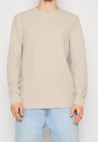 Långärmad beige ribbad tröja med rund hals och subtil "BOSS"-logotyp. Avslutad med en texturerad tyg och kontrasterande sömmar.