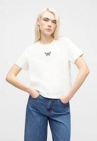 Νεαρό άτομο με ξανθά μαλλιά φοράει λευκό μπλουζάκι με μικρό μαύρο σχέδιο πεταλούδας και μπλε ψηλόμεσα τζιν, με τα χέρια στις τσέπες.