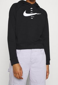 Schwarze kurze Kapuzenjacke aus weichem Stoff, mit einem weißen Nike-Logo und dem Text "Just Do It". Lange Ärmel, Design mit Kängurutasche.